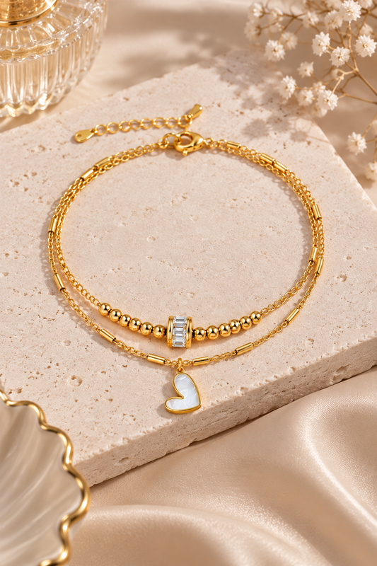 A double-strand gold bracelet with a textured chain, a beaded section, a small clear rectangular bead, and a white heart-shaped charm, displayed on a beige stone slab with satin fabric and dried flowers.